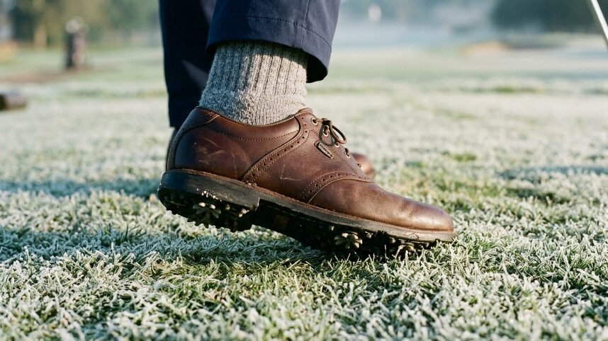 Close-up of a person wearing a brown leather golf shoe with metal spikes standing on frozen, frost-covered grass.