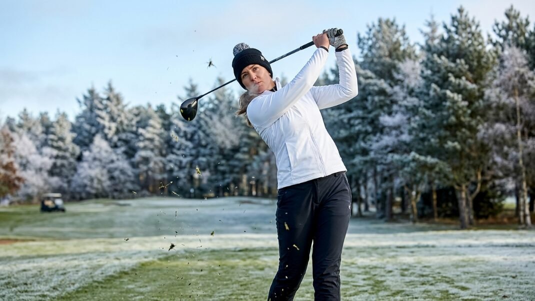 Golfer in white jacket and beanie swinging a driver, spraying grass on a cold, frost-covered winter golf course.
