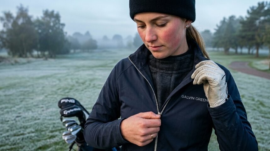 Golfer in a beanie and windbreaker standing on a frosty morning course with clubs in the background.