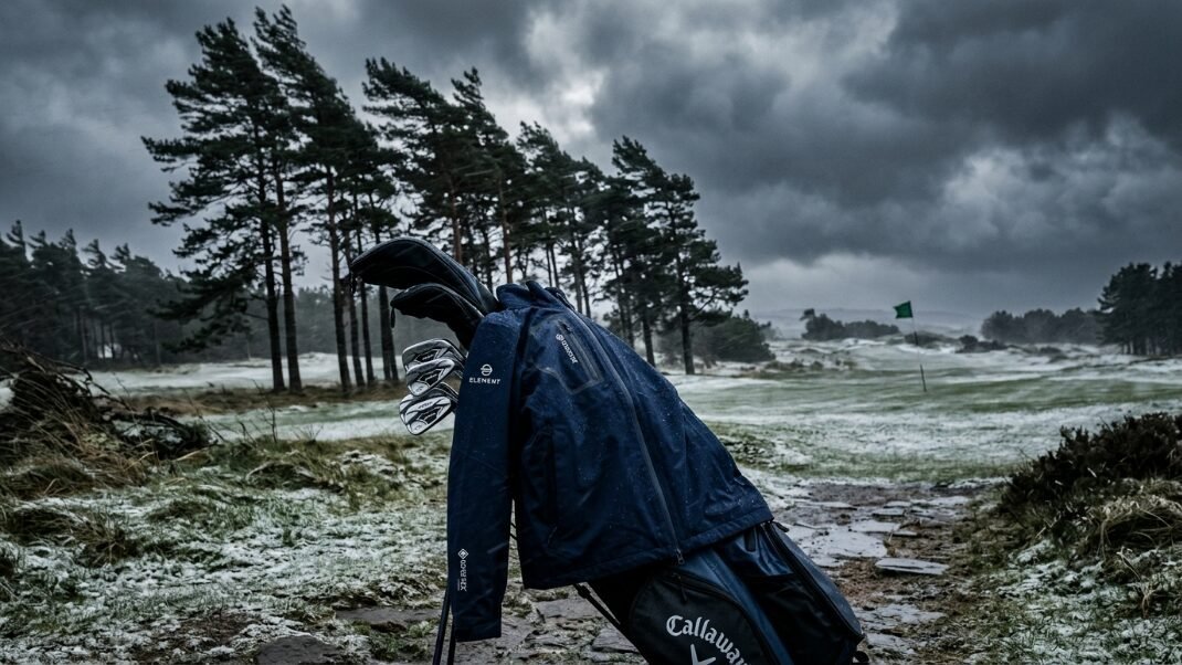 A golf bag with a jacket draped over it on a snowy, windy fairway under dark, dramatic storm clouds.