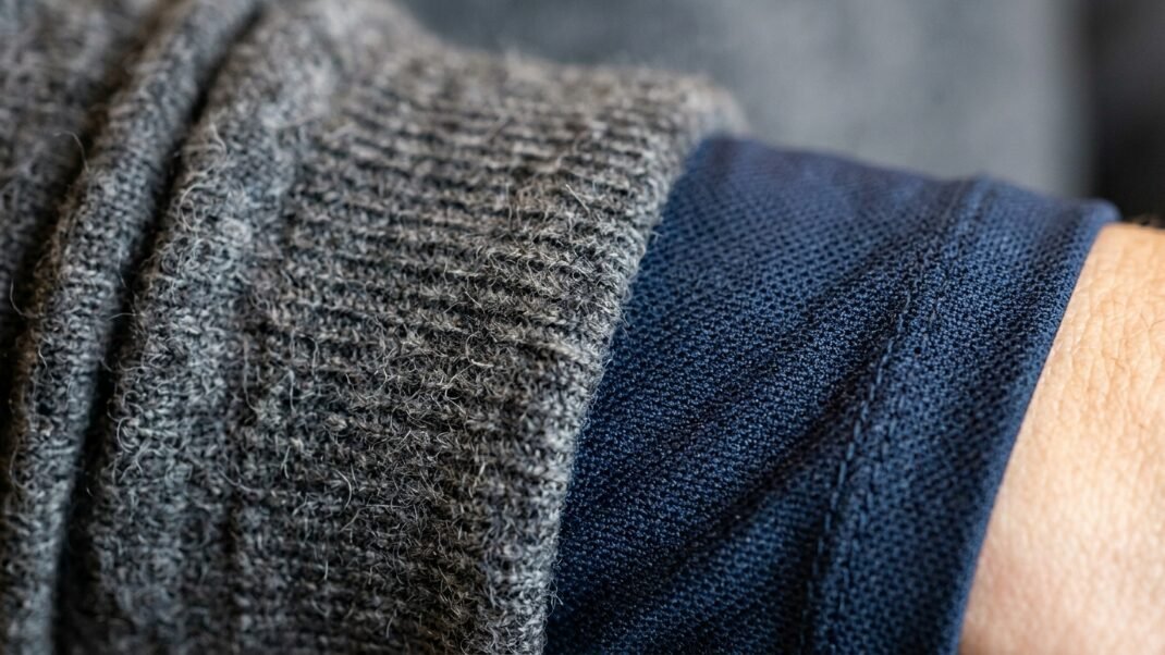 Close up of a fuzzy grey knit sweater sleeve layered over a dark blue polo shirt on a person's arm.