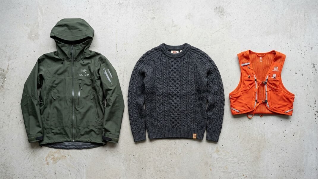 Top-down view of an Arc'teryx shell, a dark grey cable knit sweater, and a bright orange Salomon hydration vest.