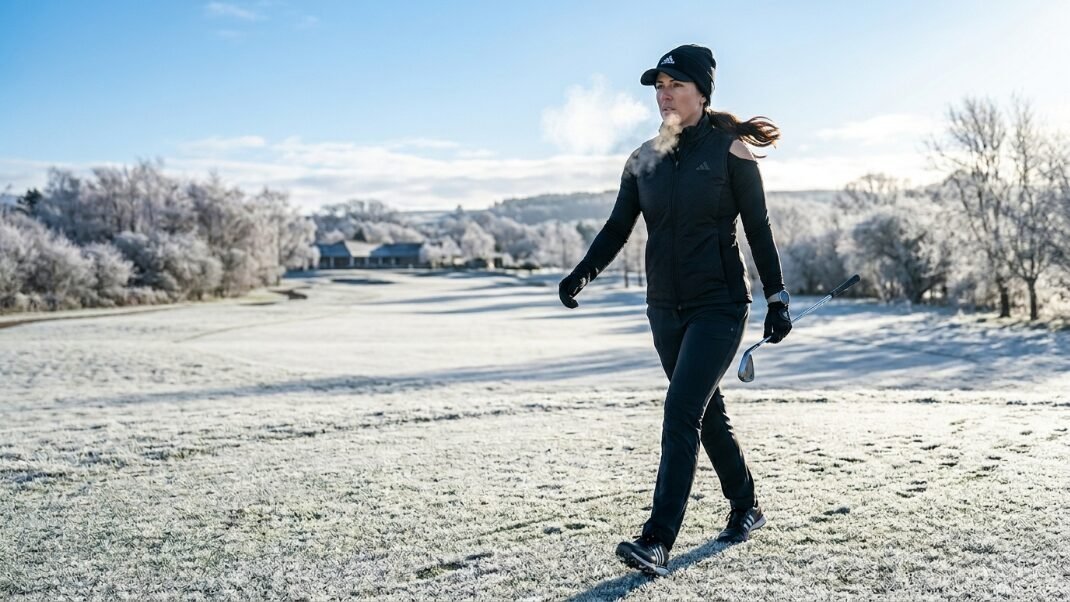 A woman in winter athletic gear exhaling visible breath while walking with a golf club on a frost-covered fairway.