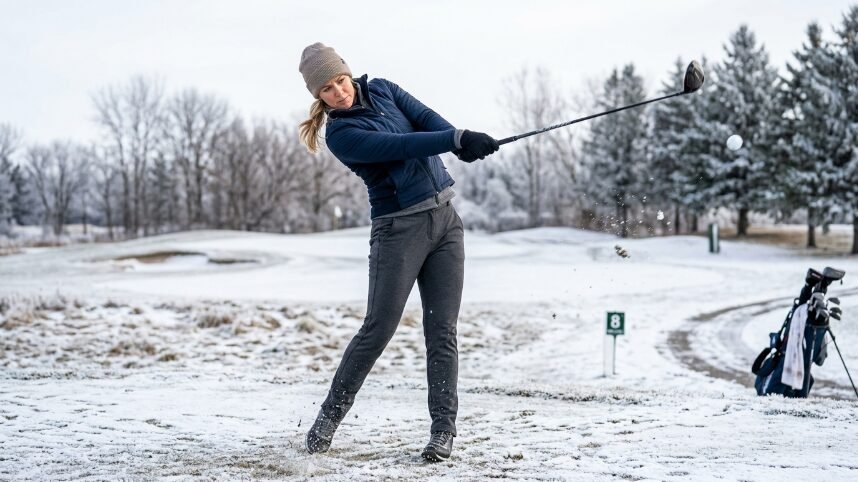 A woman wearing a winter hat and vest follows through after hitting a golf ball on a frosty, snow-dusted fairway.
