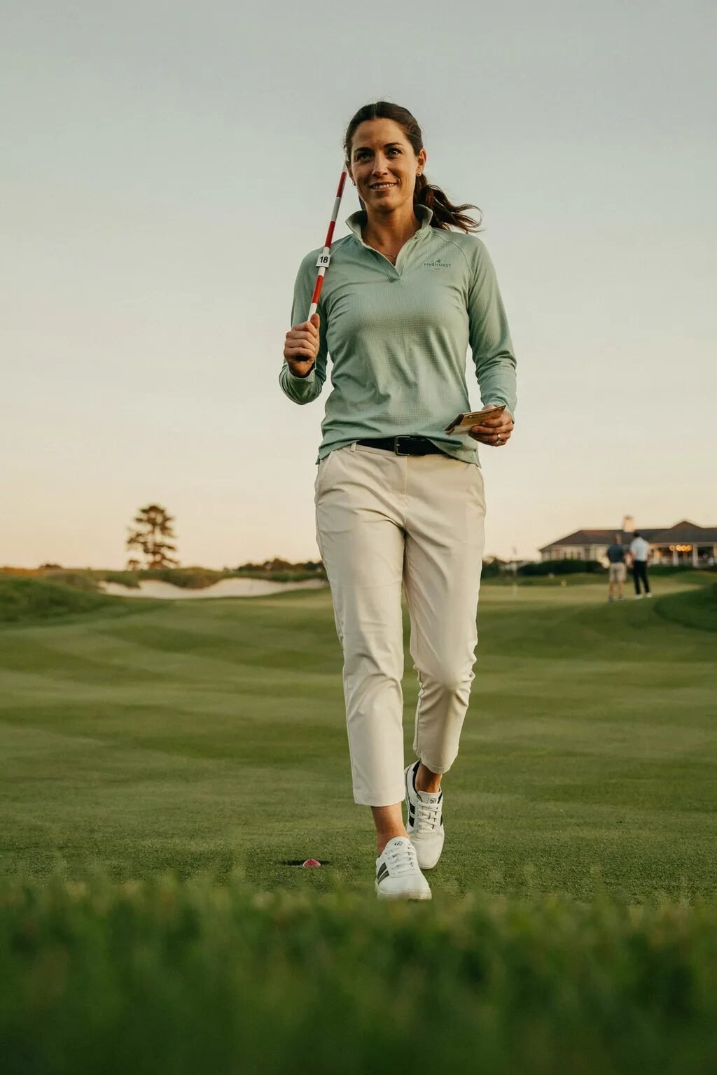 Smiling female golfer in a light green shirt and beige pants walks on a golf course with the number 18 pin flag.