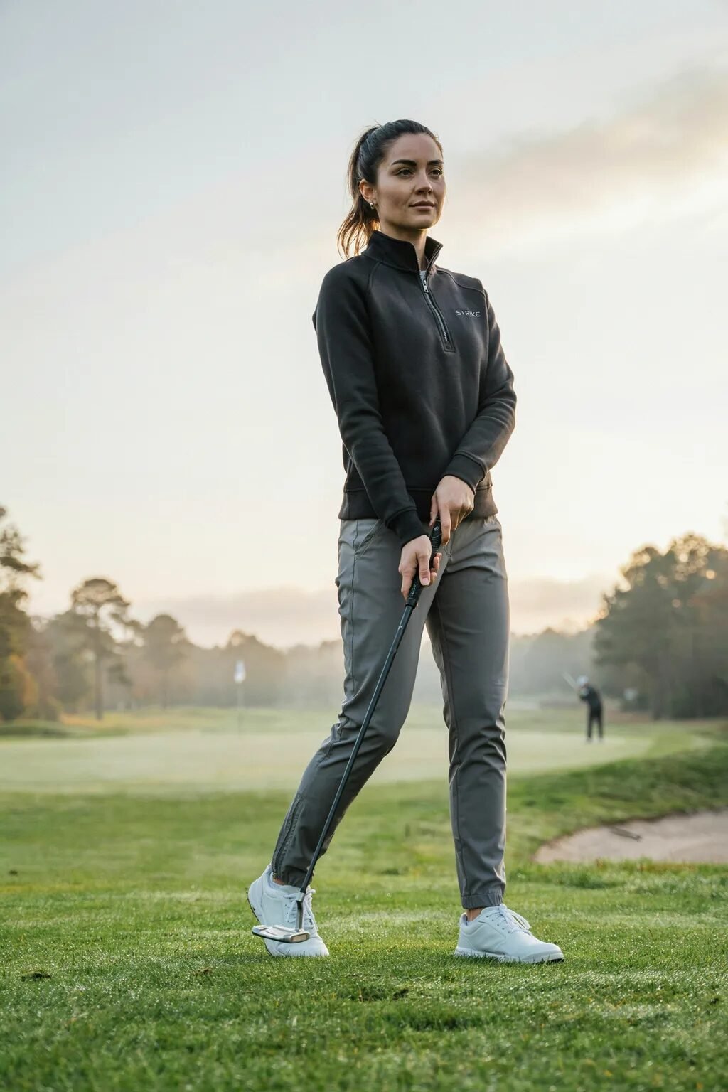 Woman in a black pullover and grey trousers stands on a dewy golf course holding a putter.
