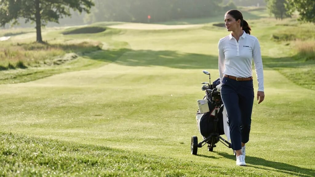 Professional female golfer pulling a push cart with clubs on a bright, grassy golf course fairway.