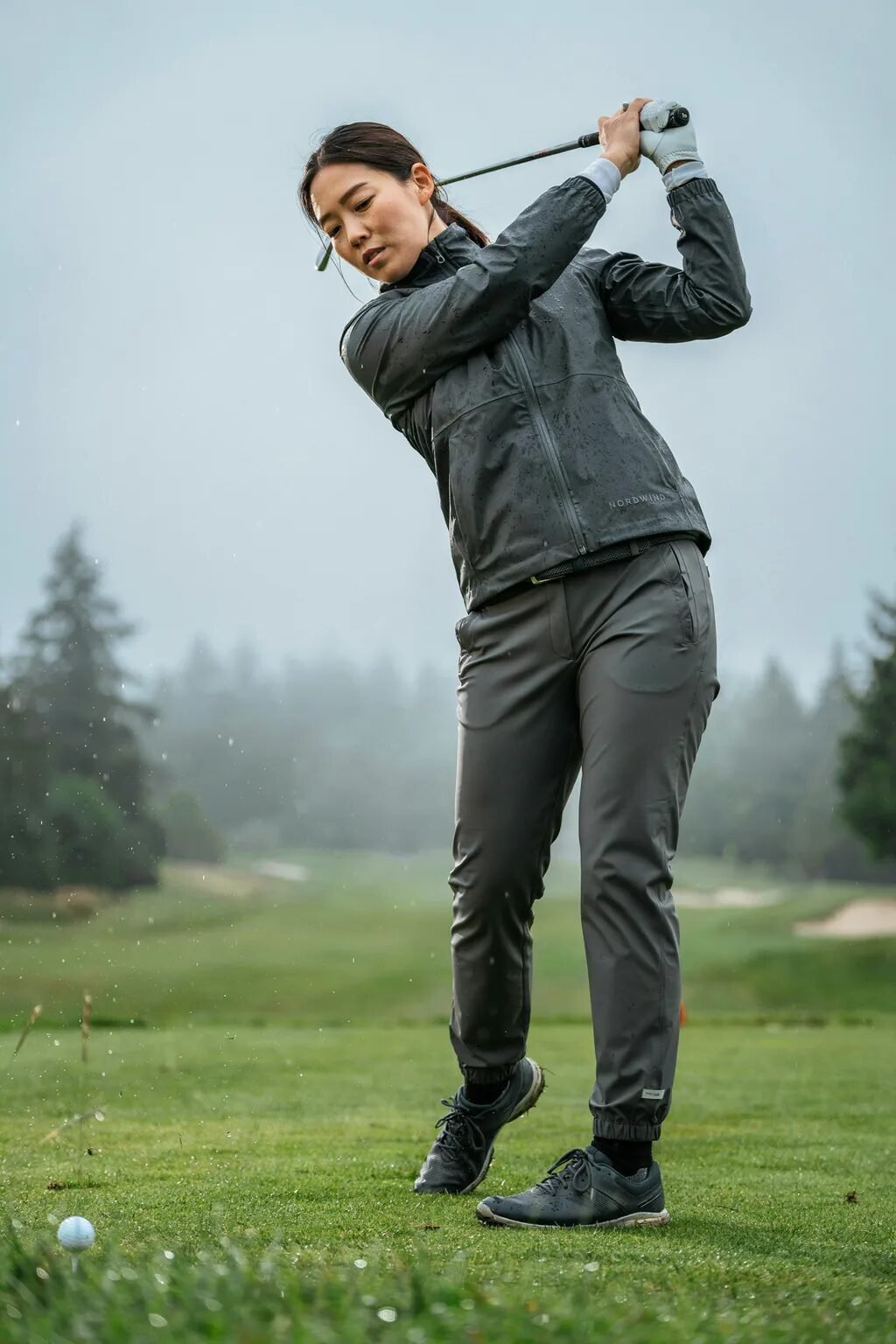 A woman wearing a dark grey waterproof jacket and pants hits a golf ball during a rainy day on the course.