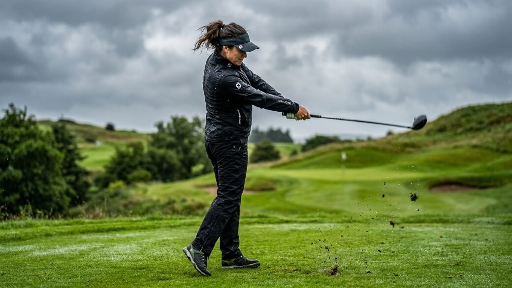 A woman in rain gear follows through on her golf swing on a lush green course under overcast skies.