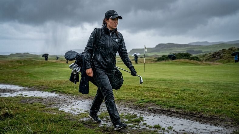 Woman wearing waterproof rain gear carrying golf clubs while walking across a muddy path in stormy weather.
