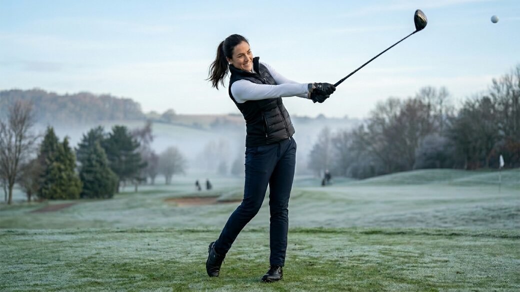 Female golfer in a black vest and white shirt hitting a ball on a frosty, misty golf course.