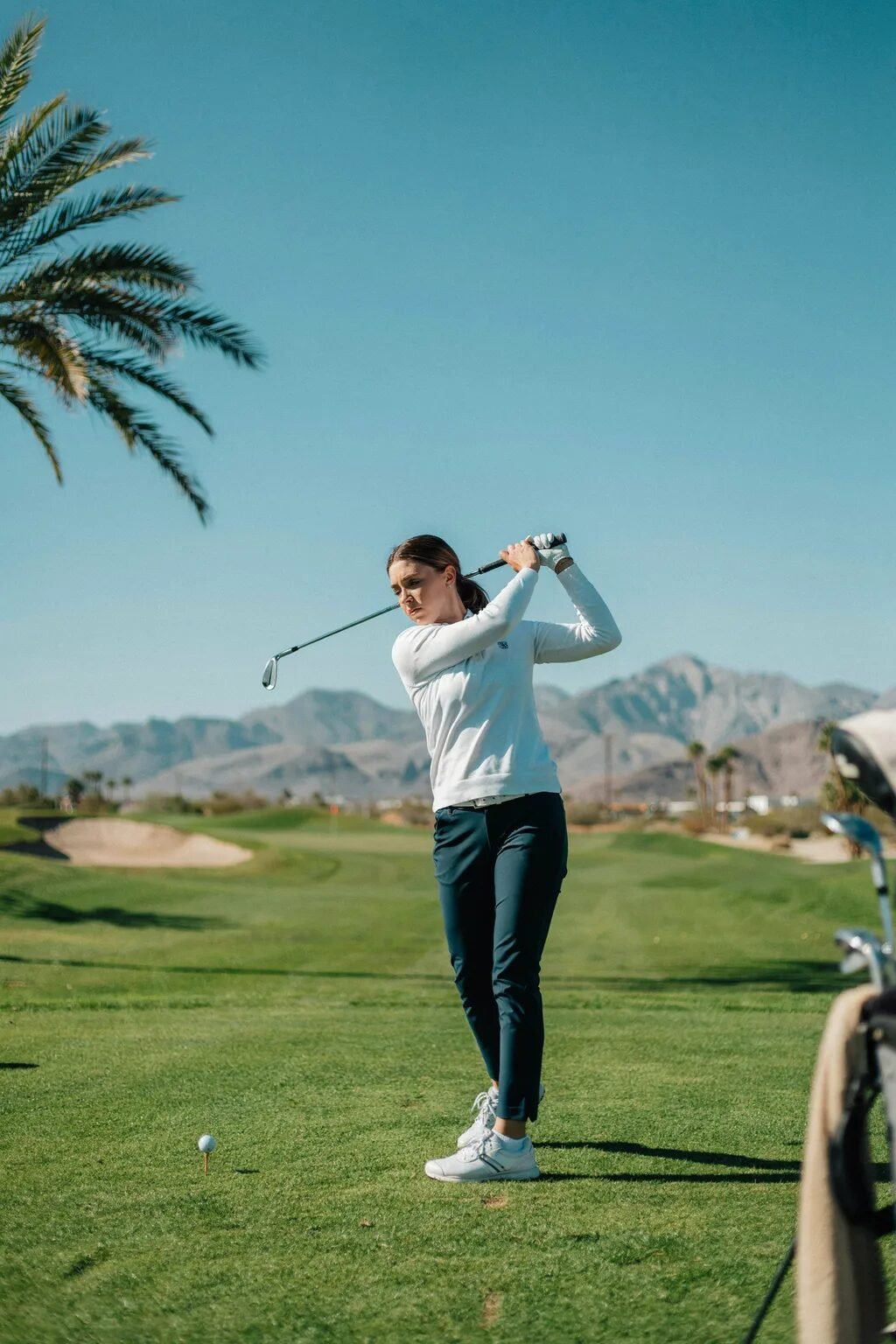 Golfer in white long-sleeved shirt and blue pants completing her follow-through on a sunny course with mountains.