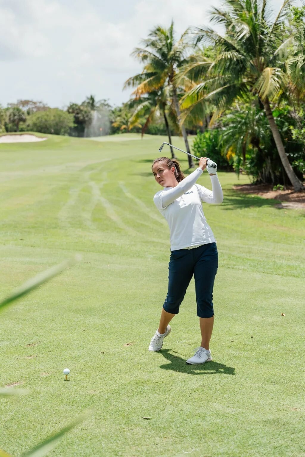 Golfer finishing her swing on a sunny green course with palm trees in the background.