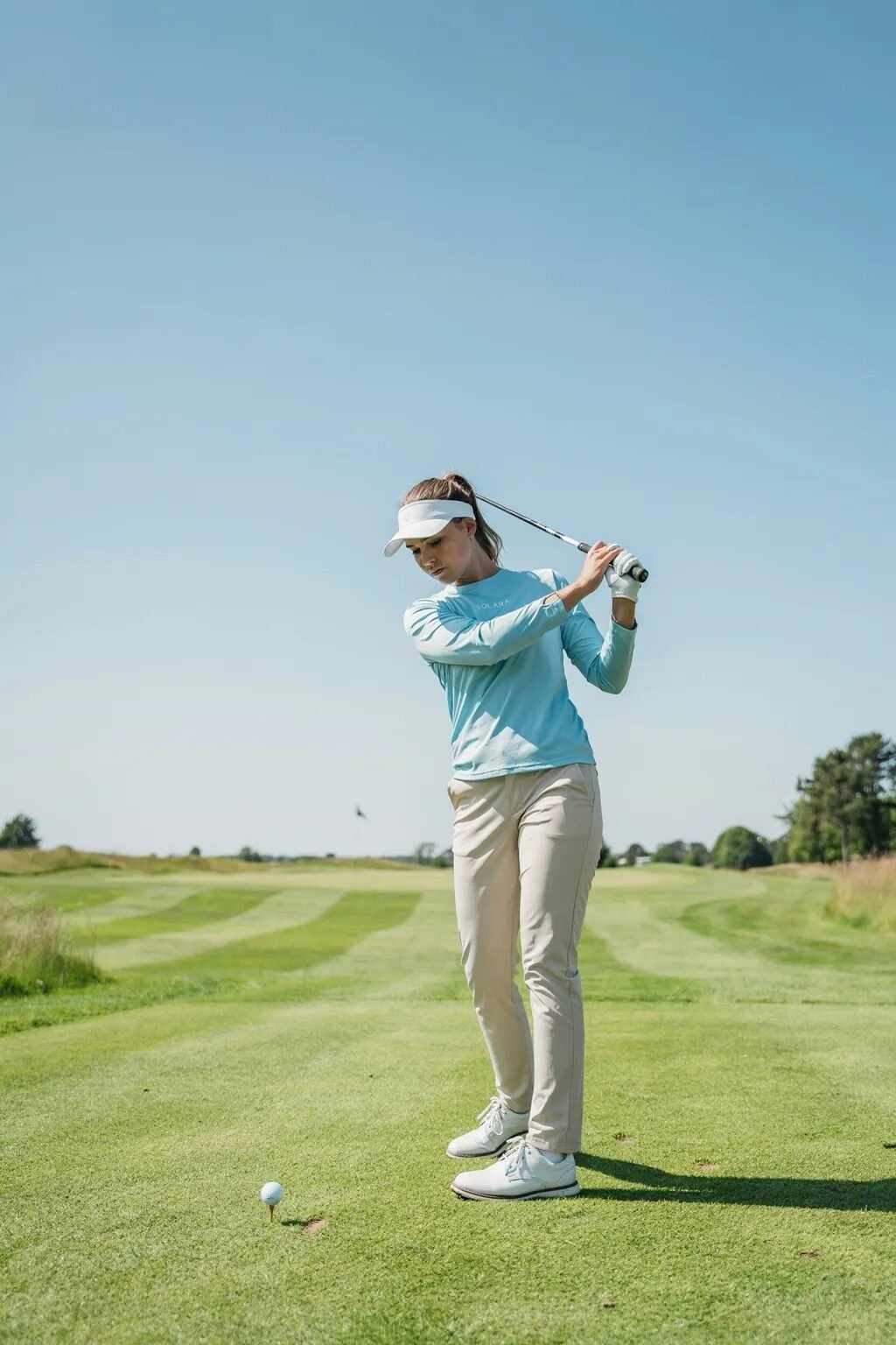 Female athlete wearing a visor and light blue shirt swinging a golf club on a sunny day.