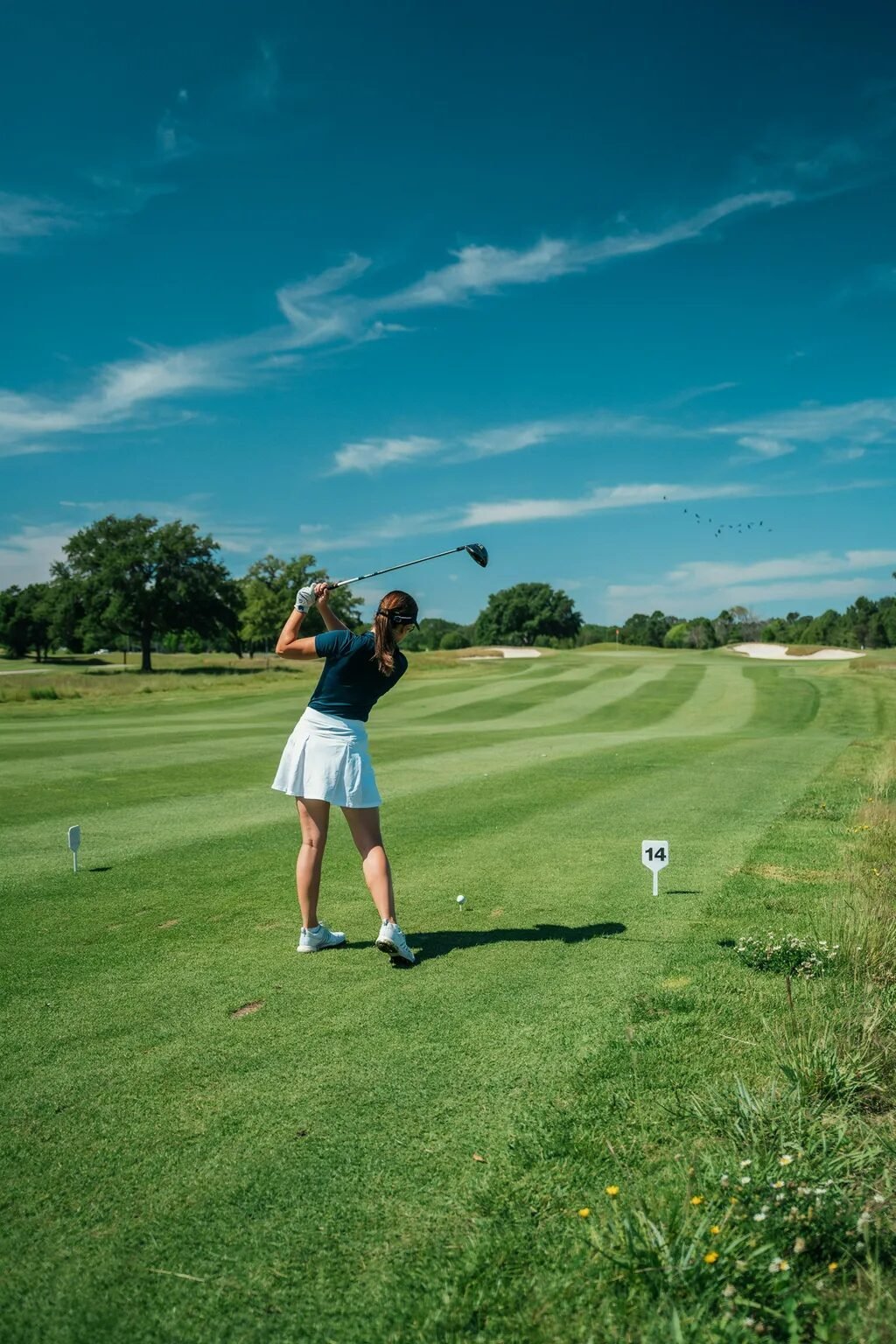 Golfer in a dark shirt and white skirt hitting a ball on a sunny golf course with a number fourteen tee sign.