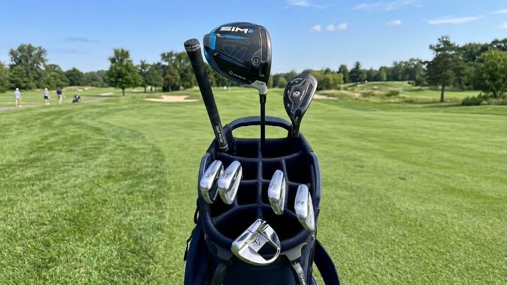 Close up of a blue golf bag with irons, a driver, and a putter set against a green fairway background.