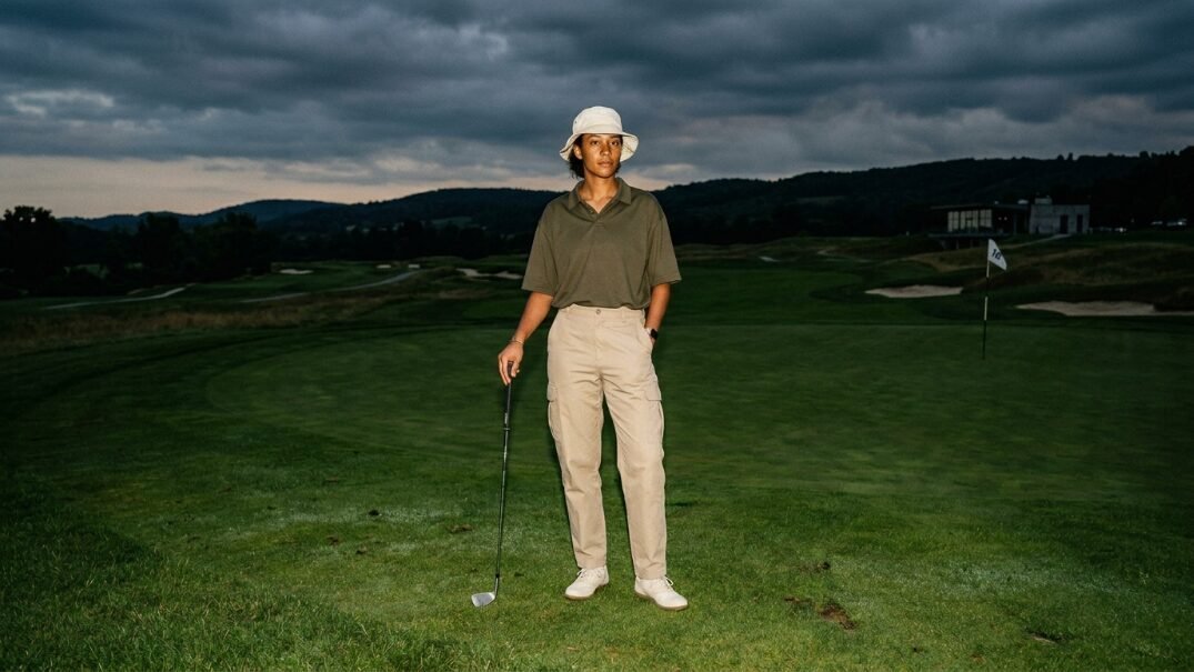 A woman in a bucket hat and polo shirt stands on a golf green holding a club under a dramatic cloudy sky.