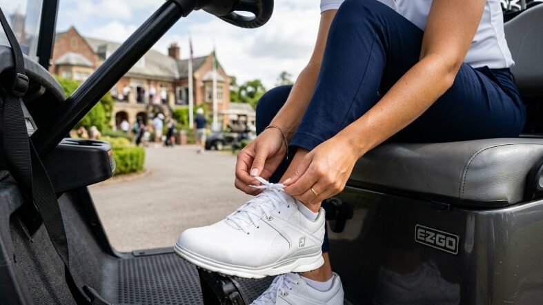 Close up of a female golfer wearing navy pants tying the laces on her white FootJoy golf shoes.