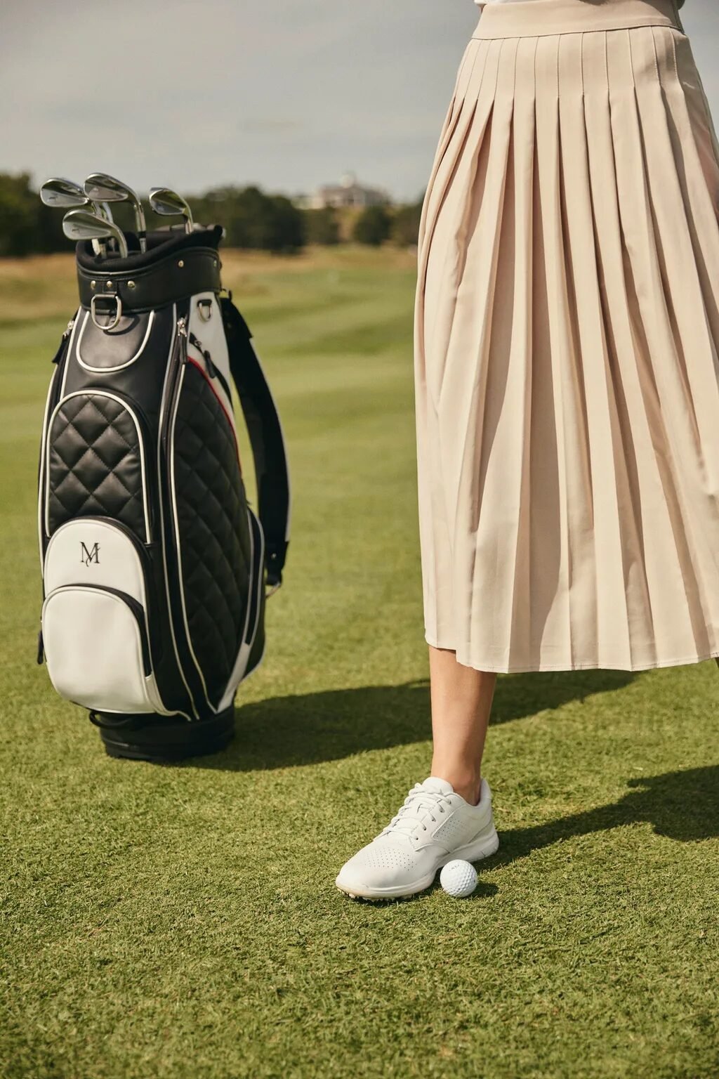 Close up of a female golfer wearing a beige skirt and white shoes next to a black and white quilted golf bag.