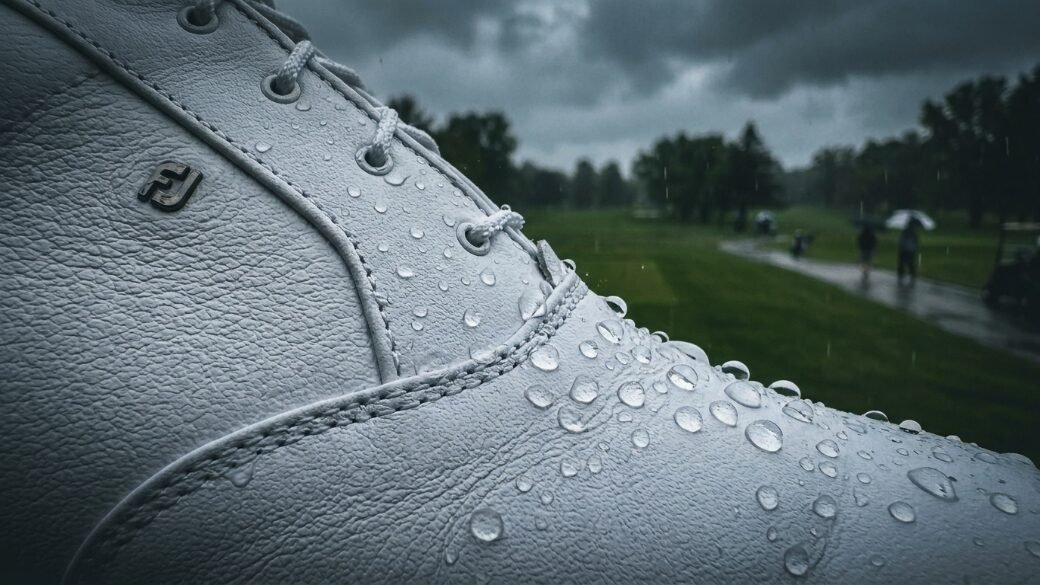Wet white leather shoe with FJ logo covered in raindrops on a rainy golf course.