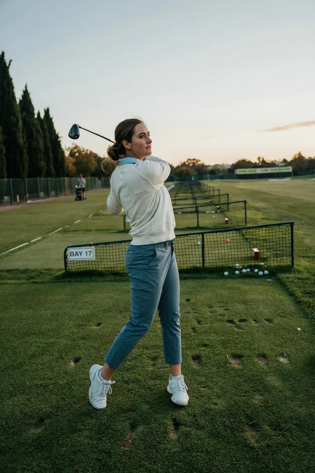 A female golfer in a beige sweater and grey pants completes her golf swing at a driving range.