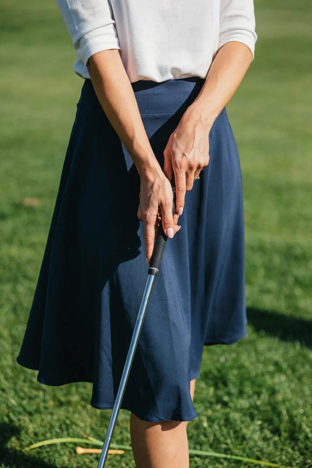 Close up of a woman wearing a white shirt and dark blue skirt standing on a grass golf course with her golf club.