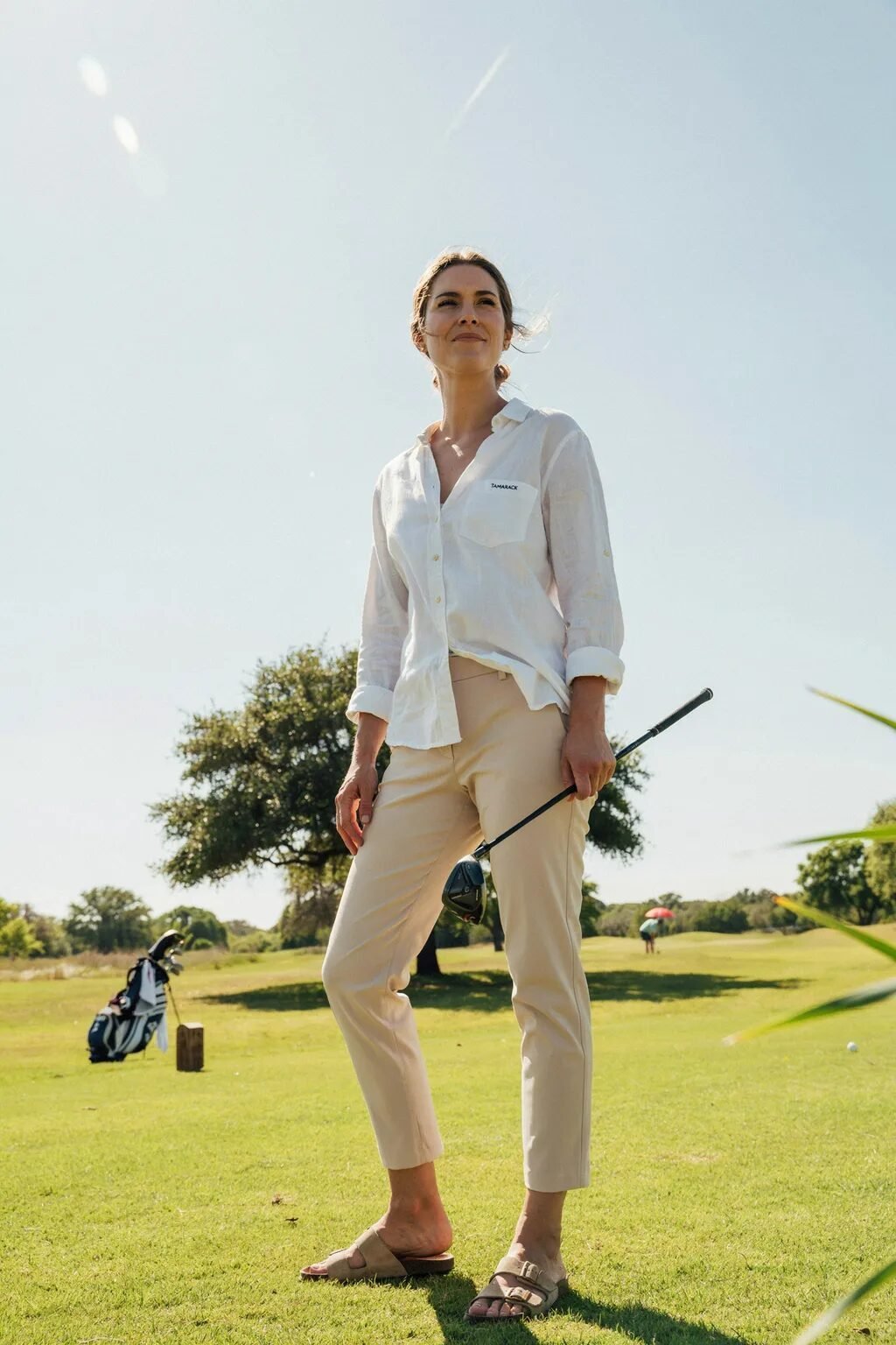 A woman in a white linen shirt and khaki pants poses with a golf club on a green course under a bright sky.