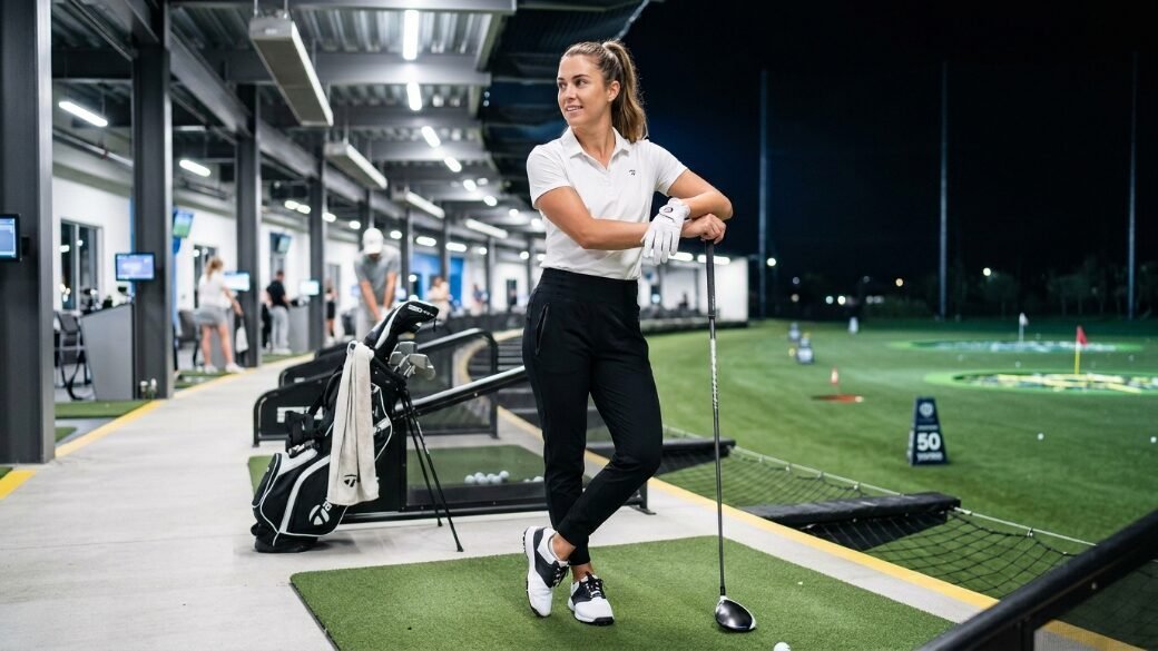 Woman in a white polo and black pants poses with a golf club at a nighttime driving range.