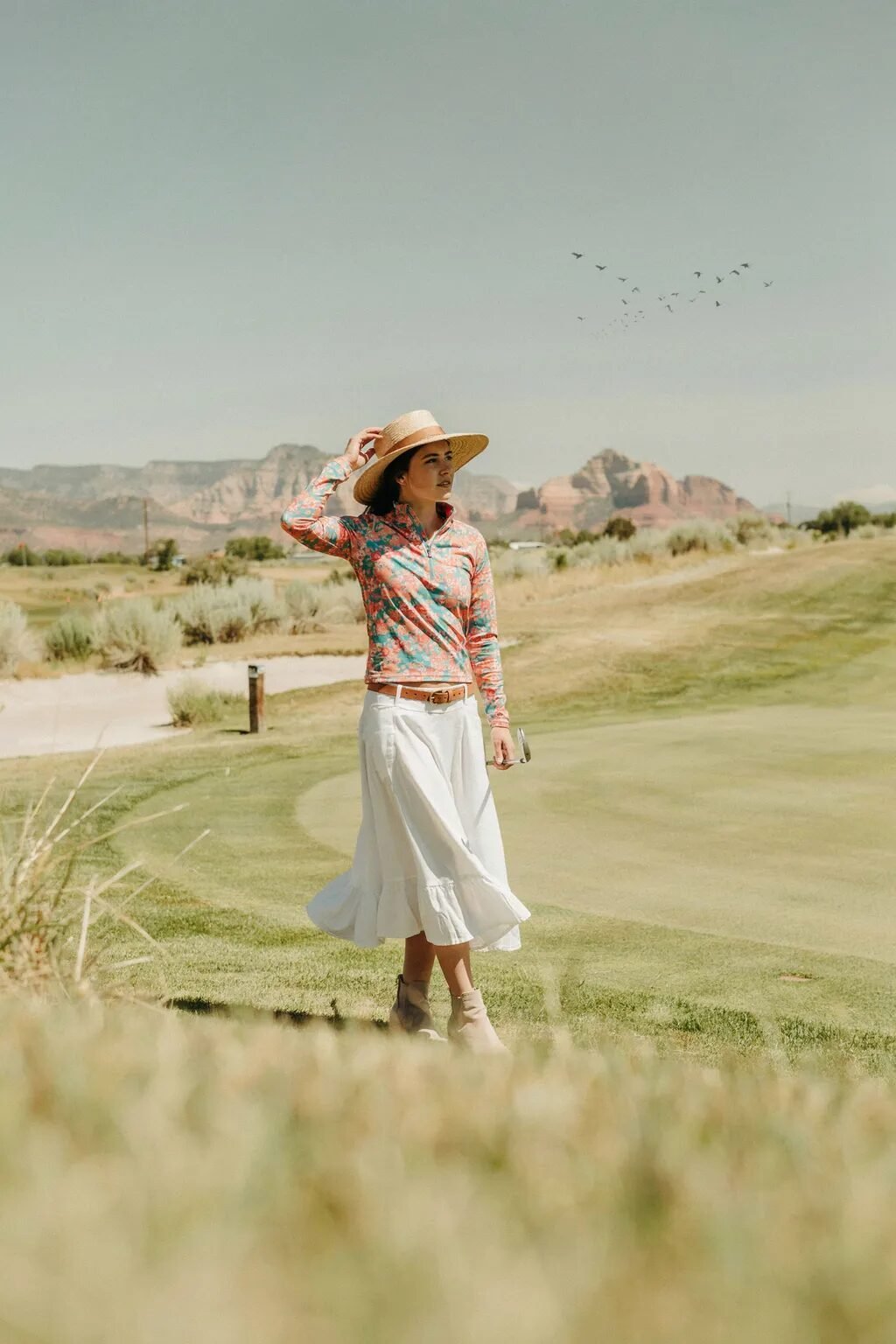 Golfer wearing a sun hat and white skirt walking across a desert course with red rocks in the background.