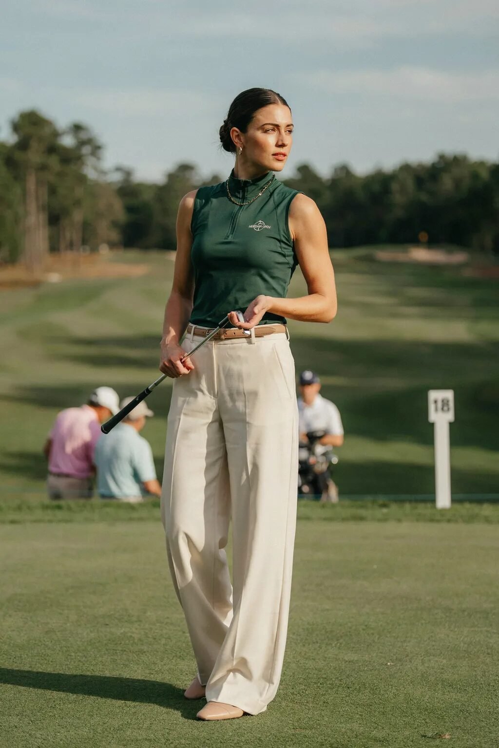 A woman in a sleeveless dark green golf shirt and wide-leg cream pants holding a golf club on a course.