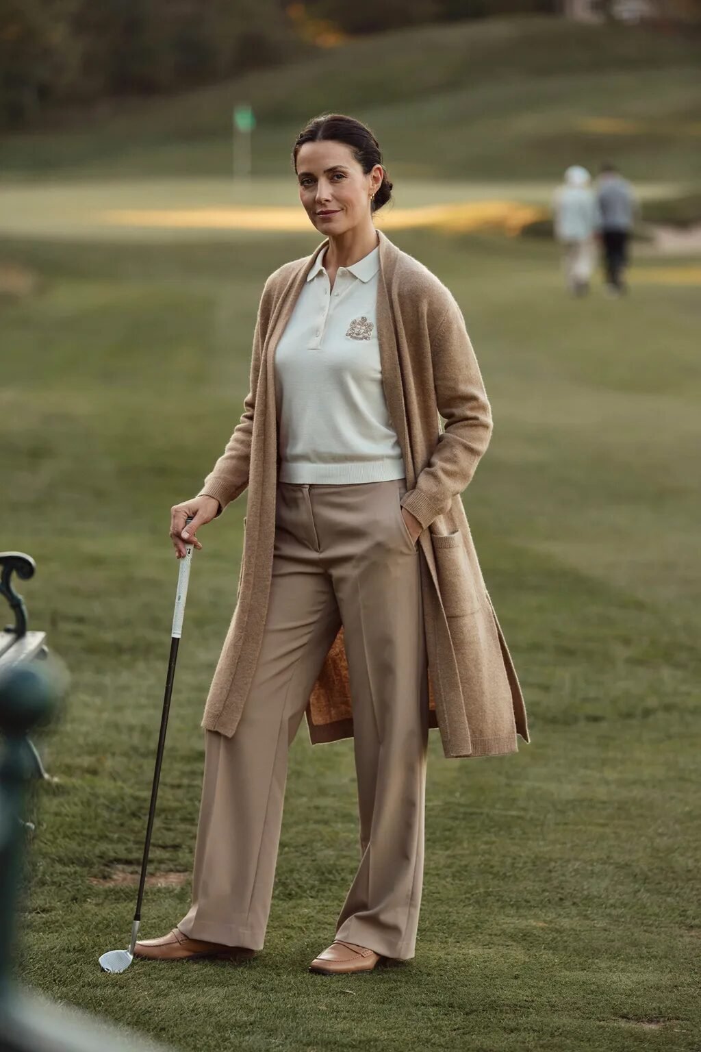 A woman in a cream polo and long beige sweater holds a golf club while standing on a grassy fairway.