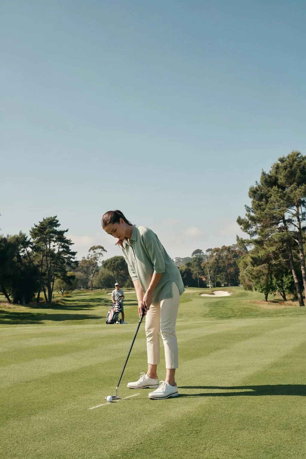 Female golfer in a green polo shirt and white pants preparing to strike a golf ball on a lush putting green.
