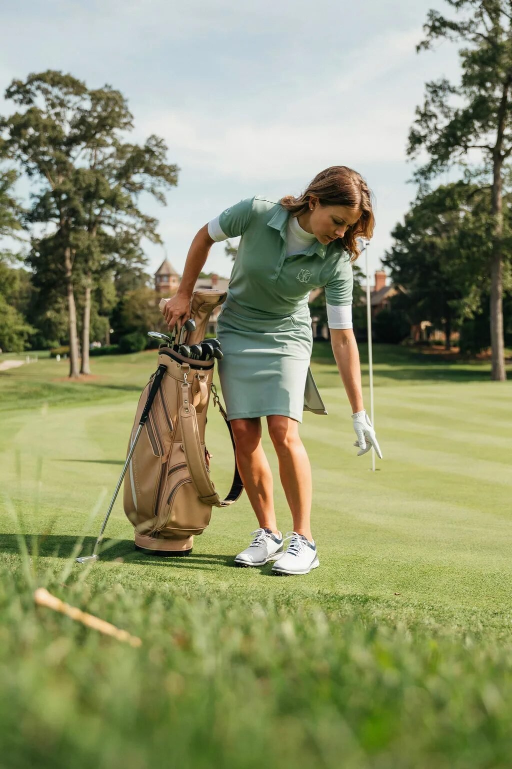 A woman in a sage green outfit standing next to her golf bag on the grass.
