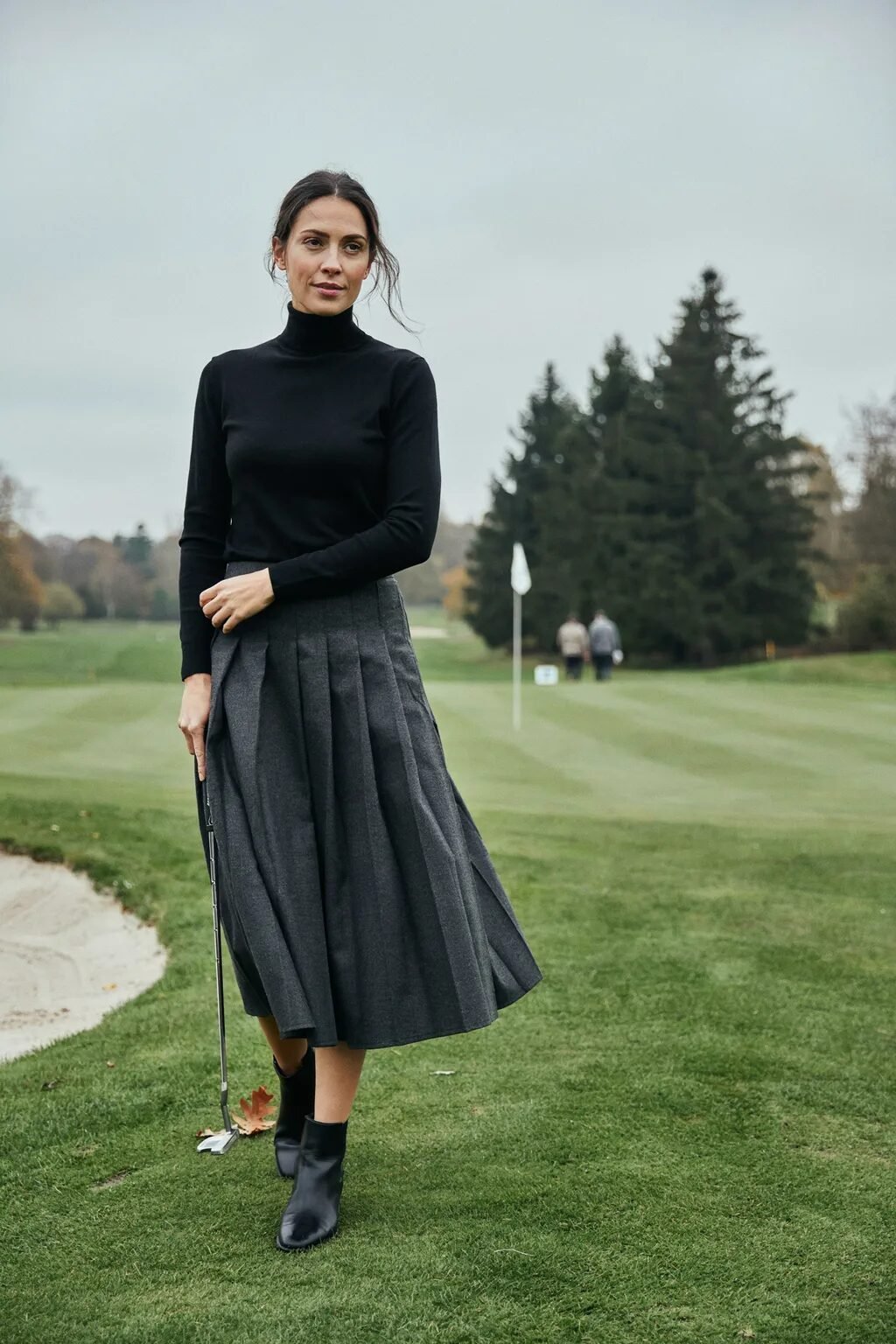 A woman in a black turtleneck and grey skirt holding a golf club on a grassy course with trees in the background.