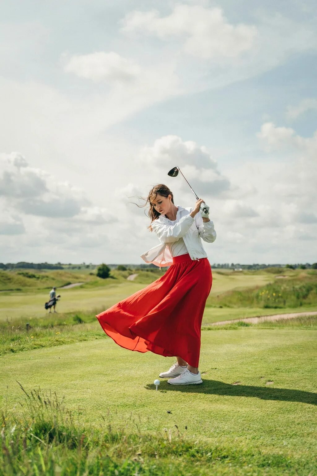 Young woman wearing a white jacket and red maxi skirt swinging a golf club on a sunny course.
