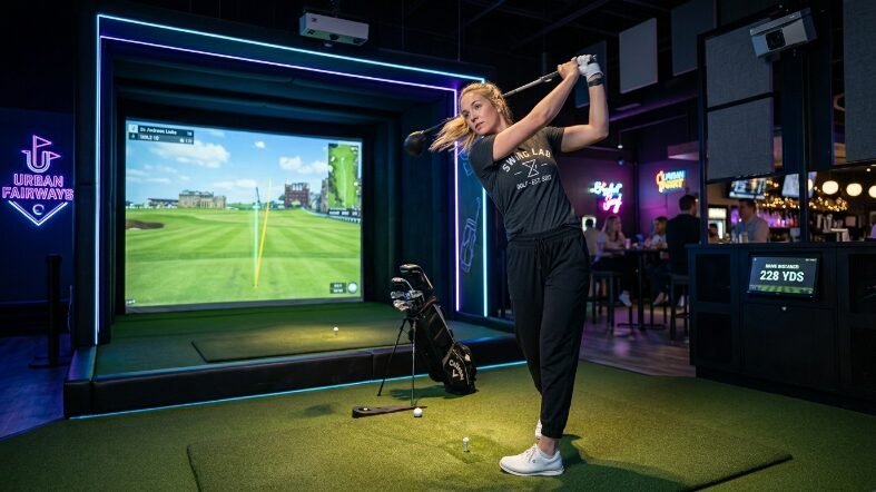 A female golfer in a dark t-shirt hitting a ball inside a brightly lit digital golf entertainment center.