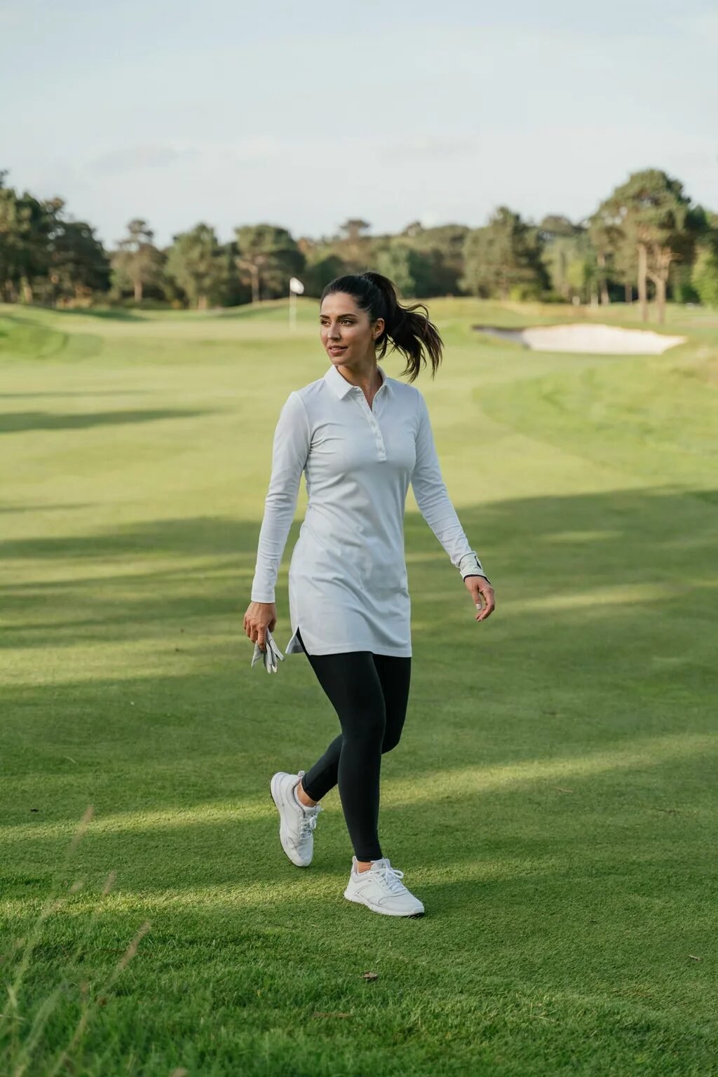 Woman in white long-sleeve polo and leggings walks across a grassy golf course holding a glove.