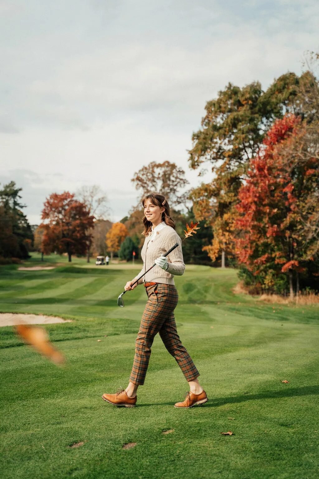 Young woman in a beige cardigan and plaid trousers walks on a lush green golf course with fall foliage.