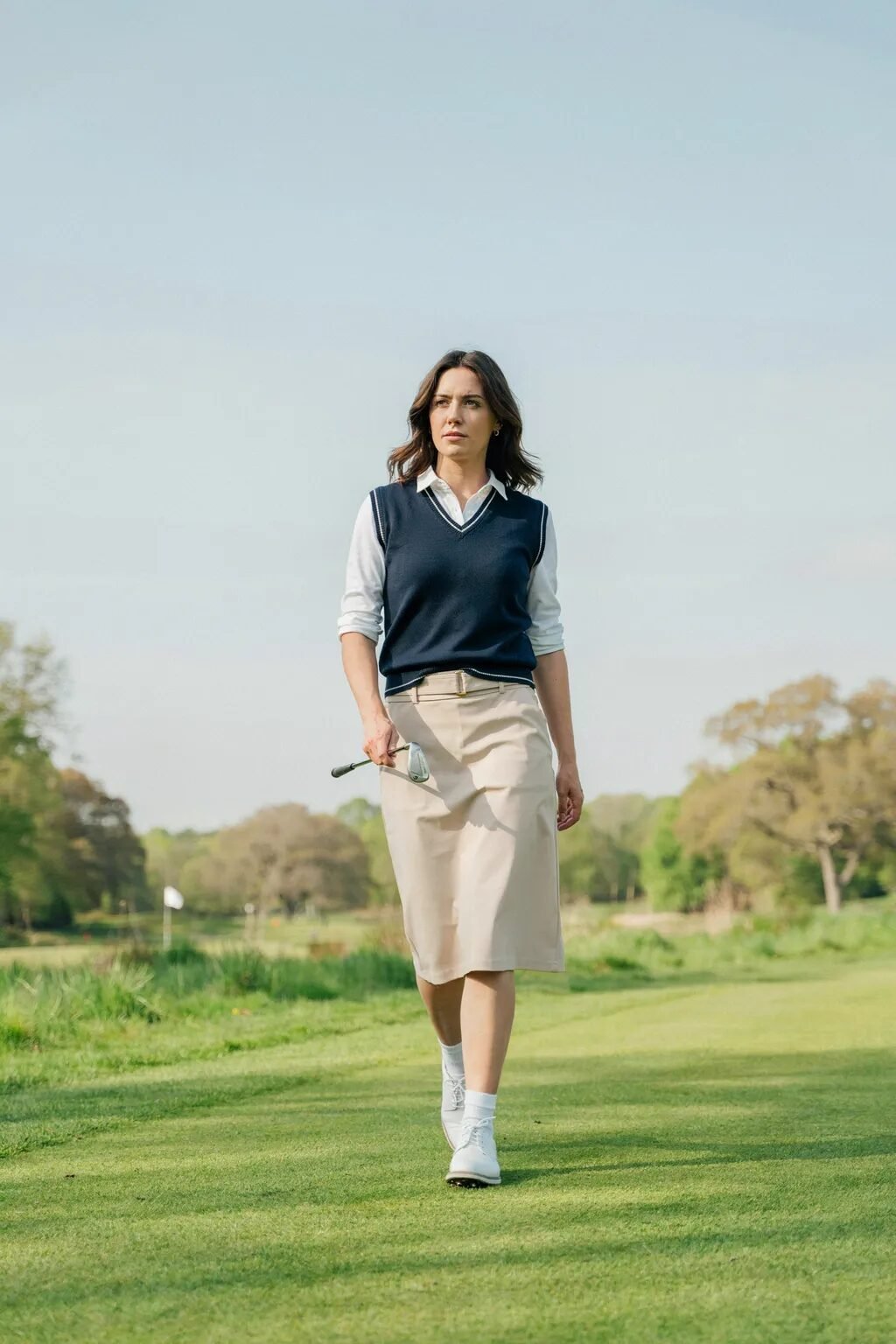 Woman in navy sweater vest and khaki skirt walking across a green golf course carrying a club.