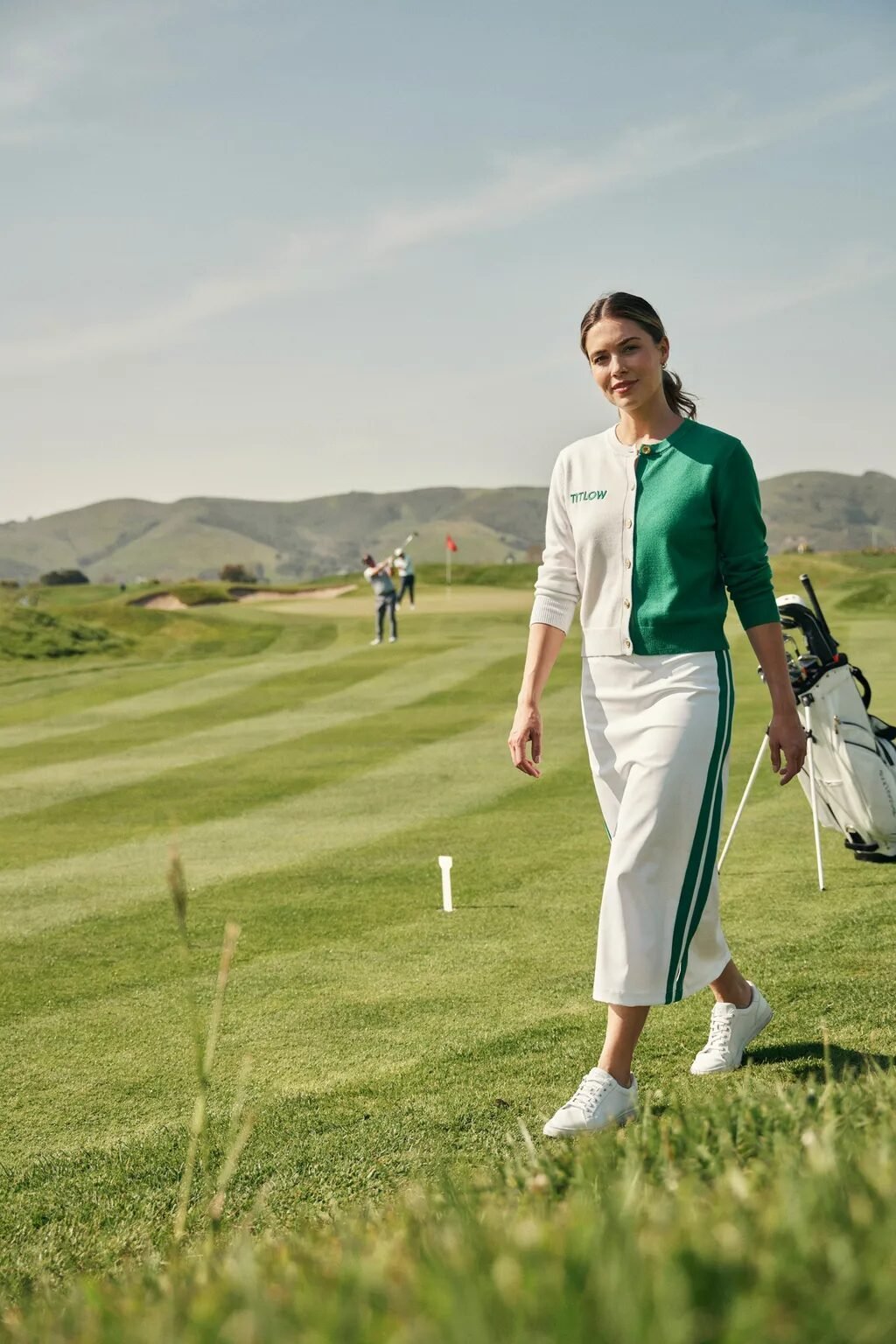 A woman wearing a green and white cardigan walks on a green golf course with a bag of clubs nearby.
