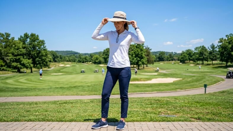 A woman in a white long-sleeve shirt and blue trousers stands on a golf course path with a beige sun hat.