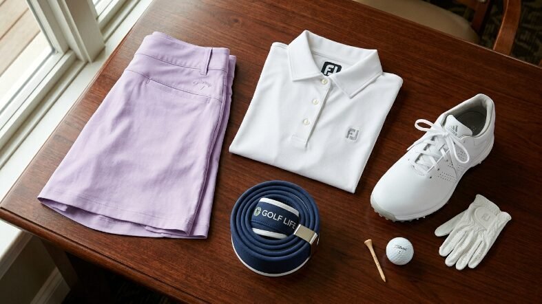 Lavender shorts, white polo, golf shoe, glove, belt, and ball on table.